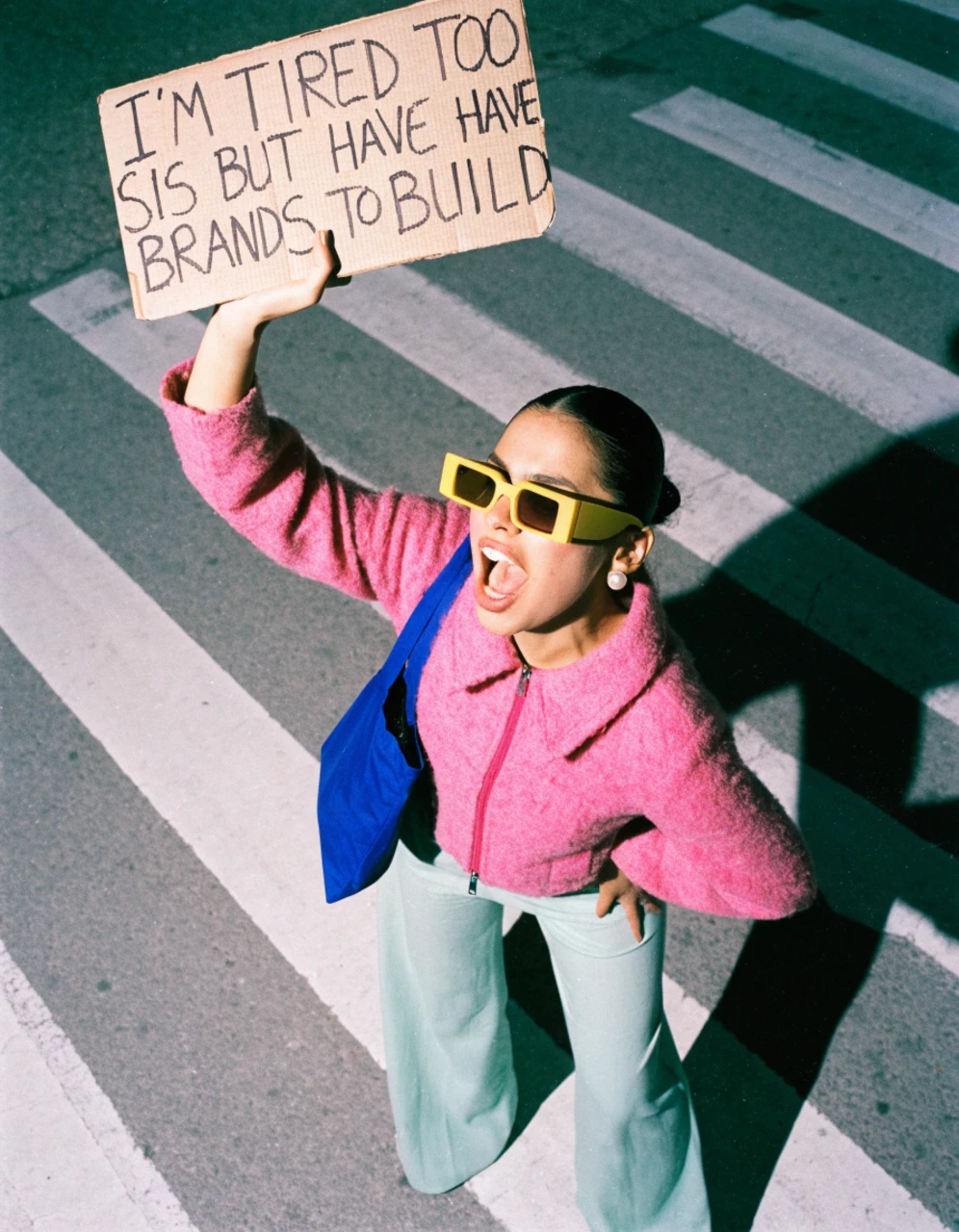 A woman in a pink jacket on a crosswalk holding up a cardboard sign that says she is tired too but has brands to build