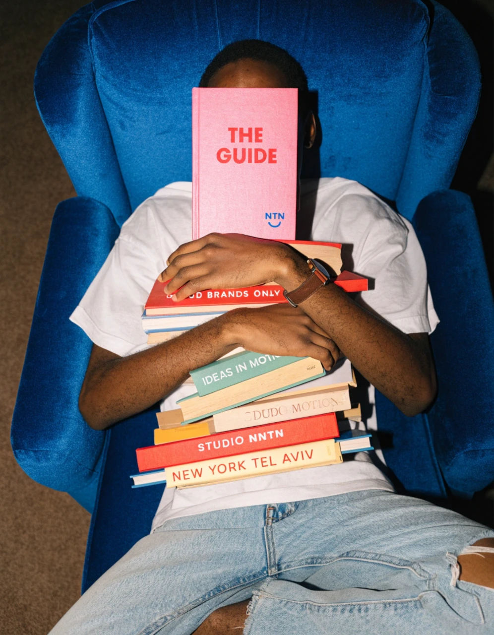 A person lying in a blue armchair, holding a stack of books with a pink copy of The guide over their face